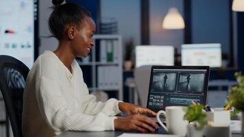 Black delighted videographer editing movie on professional laptop sitting at desk in business office at midnight. Creative editor working at night at new project processing audio film montage. photo