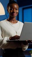 Black business woman looking at camera smiling holding laptop standing near desk in start-up company late at night. Focused employee using technology network wireless doing overtime for job photo