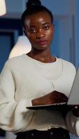 Exhausted black businesswoman holding laptop typing and sighing looking at camera standing in start-up company late at night. Focused employee doing overtime for job respecting deadline of project photo