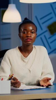 Pov of african businesswoman having conference with team during midnight looking in camera in workplace. Freelancer using wireless technology network talking on virtual meeting doing overtime photo