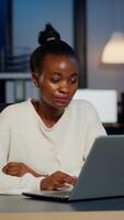 Stressed african manager woman working with financial graphs checking graphs, typing on laptop, reading raports late at night in start-up office doing overtime to respect deadline of project. photo