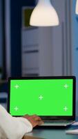 Back view of black business woman looking at laptop with greenscreen, green mockup, chroma key desktop sitting at desk in start up business office overworking. Freelancer watching at isolated display photo
