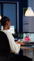 African freelancer working remotely discussing with woman partner online sitting in front of laptop in start-up office talking during virtual meeting at midnight, using headphone doing overtime photo