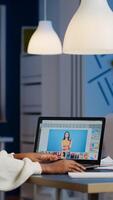 Dark skin freelancer retoucher woman working overtime on laptop with photo editing software. Professional graphic editor retouching photo of a client during night time in home office on performance pc