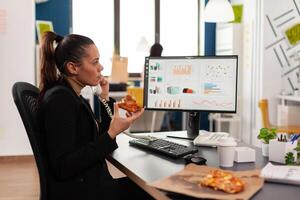 Closeup of businesswoman sitting at desk in front of computer eating pizza slice while talking at landline with remote company manager. Lunch meal break package delivered at startup office. photo