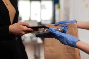 Delivery guy wearing protective gloves receiving paymant from businesswoman using smartphone nfc, technology over pos terminal service for delicious tasty meal lunch in paper bag. photo