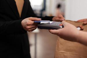 Close up of business woman holding credit card of pos, using contactless technology. Customer paying for their order with a credit card. Holding takeaway food in paper bag. photo
