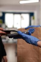 Delivery guy wearing protective gloves receiving paymant from businesswoman using smartphone nfc, technology over pos terminal service for delicious tasty meal lunch in paper bag. photo