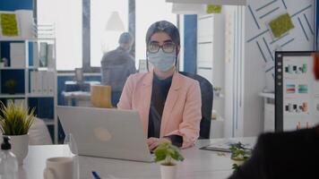 Portrait of business woman with protection face mask working on laptop computer in office during coronavirus pandemic. Coworkers keeping ing ing social distancing to prevent infection with covid19 virus photo