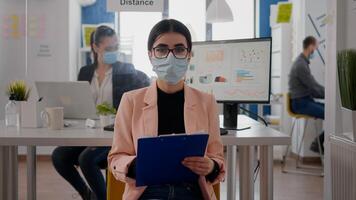 Front view of businesswoman wearing face mask while talking on online call meeting, the team working in background, keeping social distancing to avoid infection with covid19. Zoom conference teamwork photo