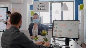 Freelancer with face mask working at financial graphics on pc sitting in business office. Coworkers talking in background about marketing mantain social distancing to prevent infection with covid19 photo