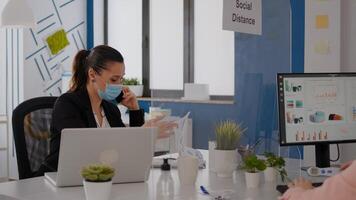 Woman with protective face mask working on laptop computer in company office checking statistics while talking on phone.Team workers respecting social distancing to avoid infection with covid19 virus photo