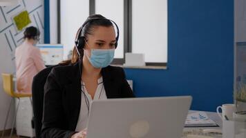 Executive manager wearing protective face mask and headphone talking with team into microphone working at business project. Businesswoman sitting in new normal office during coronavirus epidemic photo