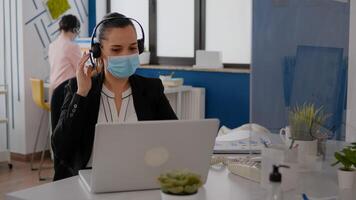 Close up of businesswoman with face mask wearing headset talking into microphone while typing marketing statistics on laptop computer. Freelancer sitting at desk in office during covid19 pandemic photo
