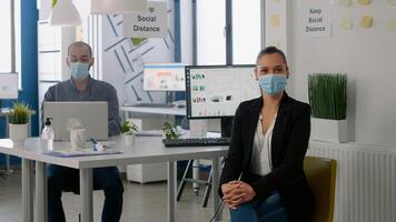 Portrait of manager woman wearing medical face mask while standing in new normal company office. Colleagues working in background for marketing project respecting social distancing to avoid covid19 photo