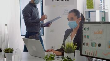 Executive manager with face mask typing marketing information on laptop computer while sitting at desk table in company office. Team respecting social distance to avoid infection with covid19 photo