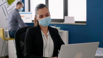 Business woman with protective face mask analysing on laptop computer marketing statistics, sitting at desk table in company office. Coworkers keeping social distancing to avoid virus disease photo