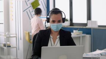 Businesswoman with protection face mask talking into microphone sitting at table desk in front of laptop computer. Coworkers keeping social distancing to prevent virus disease photo
