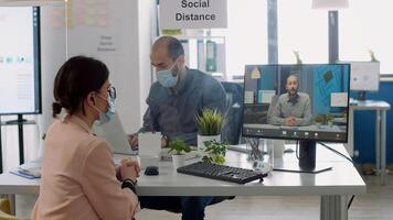 Freelancer with protective face mask talking with collegue during virtual videocall conference while sitting in business office. Coworkers respecting social distance to prevent infection with covid19 photo
