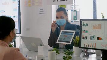 Back shot of business womens with medical face mask working together at management presentation using tablet computer while sitting in company office. Team respecting social distance photo