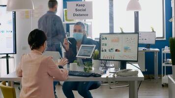 Executive manager with face mask showing statistics to her collegue using digital tablet computer. Coworkers working in new normal office keeping ing ing social distance to prevent infection with covid19 photo