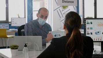 Coworkers with protective face masks talking about management statistics using tablet computer sitting in new normal company office. Team mantain social distancing to avoid infection with coronavirus photo
