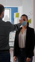 Manager with protection face mask examines collegues temperature with infrared thermometer before enter in office during coronavirus pandemic. Coworkers keeping social distance to prevent covid19 photo