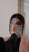 Close up of woman measuring temperature with medical thermometer to prevent covid 19. Coworkers keeping social distance wearing protection face mask at work place during coronavirus global pandemic photo