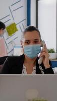 Entrepreneur with protective face mask discussing on landline while sitting at startup office desk in front of computer. Caucasian female working at business meeting during coronavirus global pandemic photo