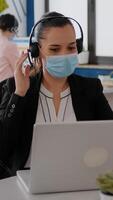 Close up of businesswoman with face mask wearing headset talking into microphone while typing marketing statistics on laptop computer. Freelancer sitting at desk in office during covid19 pandemic photo