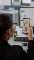 Executive business woman wearing protective face mask using phone for online videocall conference with remotely team. Coworkers working in background respecting social distance photo