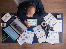 Top view of businessman using tablet pc analyzing financial charts and documents, sitting at desk in corporate office. Employee scrolling through graphs. Successful entrepreneur analyzing strategy. photo