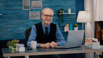 Senior man looking at camera smiling sitting in workplace at desk working from home while old wife watching tv in background. Retired business person prepared for online meeting using moden gadgets photo