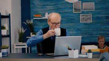 mayor empresario leyendo informes sentado en frente de ordenador portátil trabajando desde hogar Bebiendo café. retirado hombre utilizando moderno tecnología analizando mecanografía buscando mientras esposa sentado en sofá leyendo un libro foto