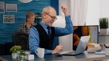 Excited senior man feel ecstatic reading great online news on laptop working from home. Retired employee using modern technology reading typing, searching while senior wife sitting on couch photo