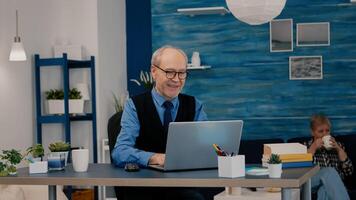Happy old manager receving good news on laptop working from home for startup company, gesturing victory. Retired employee using modern technology reading typing searching while senior wife watching TV photo