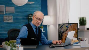Senior man using laptop and tablet in same time working in office living room at project. Busy focused old employee writing, searching, analysing reports while elderly wife watching tv in background photo