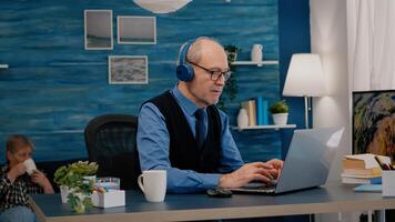 enfocado antiguo empresario con auriculares escuchando música mientras comprobación gráficos escritura en ordenador portátil trabajando desde hogar. retirado hombre utilizando moderno tecnología leyendo mecanografía, buscando en computadora foto