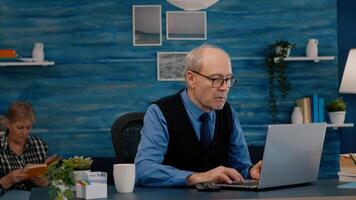 Old freelancer typing on laptop sitting in living room working remote. Busy focused elderly employee using modern technology reading typing, searching while old wife sitting on couch reading a book photo