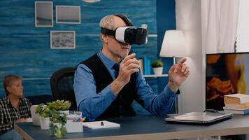 Retired man experiencing virtual reality using vr headset in living room working from home. Old remote employee searching, analysing financial reports while elderly wife watching tv in background photo