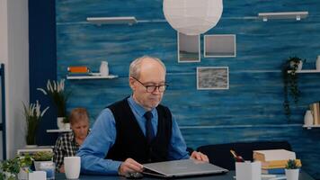 Elderly man freelancer opening laptop squinting while reading emails working from home. Retired man using modern technology reading typing searching while wife sitting on couch reading a book photo