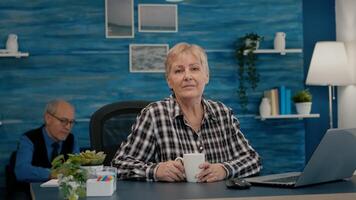 Senior focused woman looking at camera sitting in workplace at desk working from home while old wife watching tv in background. Retired business person prepared for online meeting using moden gadgets photo