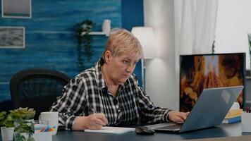 Old woman entrepreneur checking financial project of company looking on laptop analysing annual statistics data, taking notes on notebook sitting at desk in living room office working from home. photo