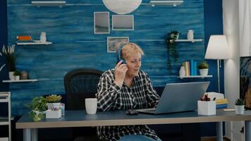 Happy old woman entrepreneur with headset listening music while checking statistics, writing on laptop working from home. Retired worker using modern technology reading typing, searching on computer photo