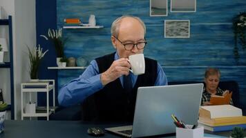 Tired elderly man sitting at desk and taking a sip of coffee, looking at laptop working from home workspace while his wife is reading a book in background. Focused old entrepreneur checking graphics photo