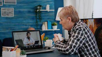 Old woman sitting at table at home looking at laptop answering question about health talks with therapist by videoconference call application, holding pills flask Remote medical consultation concept photo