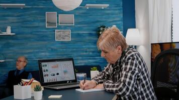 Middle aged senior older woman elearning online concept, watching business graphs, charts on laptop computer remote working, social distance learning, writing notes from home office. photo
