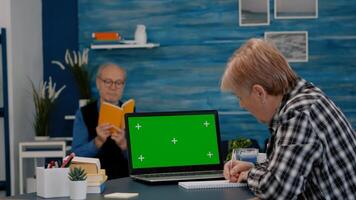 Senior woman looking at green screen monitor working from home, taking notes on notebook. Elderly businesswoman watching desktop laptop display with green mockup, chroma key, while man reading a book photo