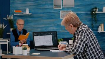 Senior woman architect analysing digital prototype with plans from laptop working from home, taking notes on notebook. Elderly designer using cad software to create a 3D concept of buildings photo