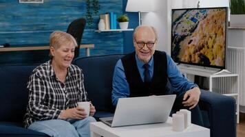 Enthusiastic couple of pensioners sitting together at home on sofa using laptop talking on call with family friends waving at camera during online meeting. Connection lifestyle concept in quarantine photo